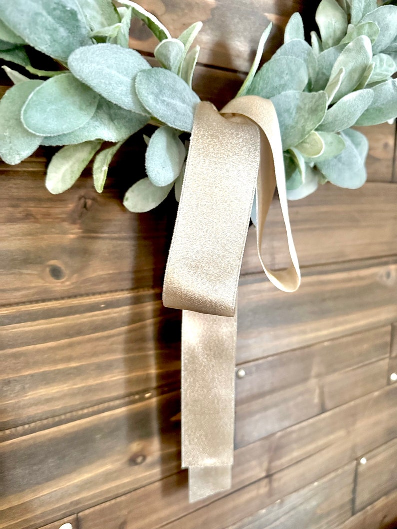 May include: A light beige ribbon with a shimmery texture hangs from a green wreath. The wreath is made of faux greenery and is hanging on a wooden wall.