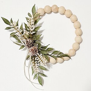 K&ouml;nnte beinhalten: Dekorativer Kranz mit einem kreisf&ouml;rmigen Design aus Holzperlen. Der Kranz ist mit gr&uuml;nen Bl&auml;ttern, wei&szlig;en Bl&uuml;ten und zwei kleinen Tannenzapfen verziert. Silberne B&auml;nder akzentuieren die floralen Elemente. Wei&szlig;er Hintergrund.