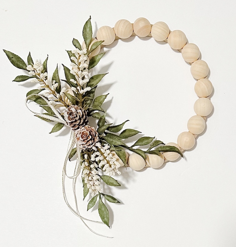K&ouml;nnte beinhalten: Dekorativer Kranz mit einem kreisf&ouml;rmigen Design aus Holzperlen. Der Kranz ist mit gr&uuml;nen Bl&auml;ttern, wei&szlig;en Bl&uuml;ten und zwei kleinen Tannenzapfen verziert. Silberne B&auml;nder akzentuieren die floralen Elemente. Wei&szlig;er Hintergrund.