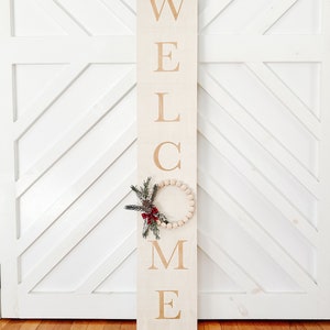 Puede incluir: Un letrero rectangular de madera con la palabra "WELCOME" en letras grandes de color marr&oacute;n claro. Una peque&ntilde;a corona redonda hecha de cuentas de madera y vegetaci&oacute;n est&aacute; unida al letrero. El letrero est&aacute; apoyado contra una puerta blanca.