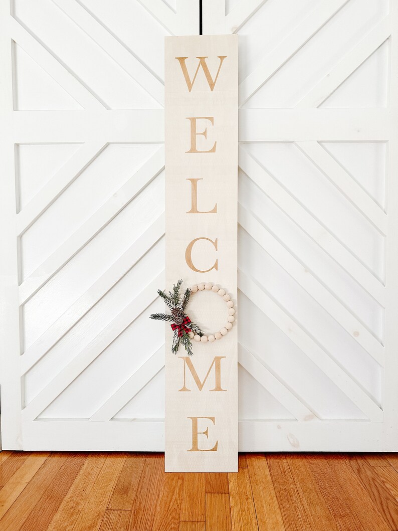 Puede incluir: Un letrero rectangular de madera con la palabra "WELCOME" en letras grandes de color marr&oacute;n claro. Una peque&ntilde;a corona redonda hecha de cuentas de madera y vegetaci&oacute;n est&aacute; unida al letrero. El letrero est&aacute; apoyado contra una puerta blanca.