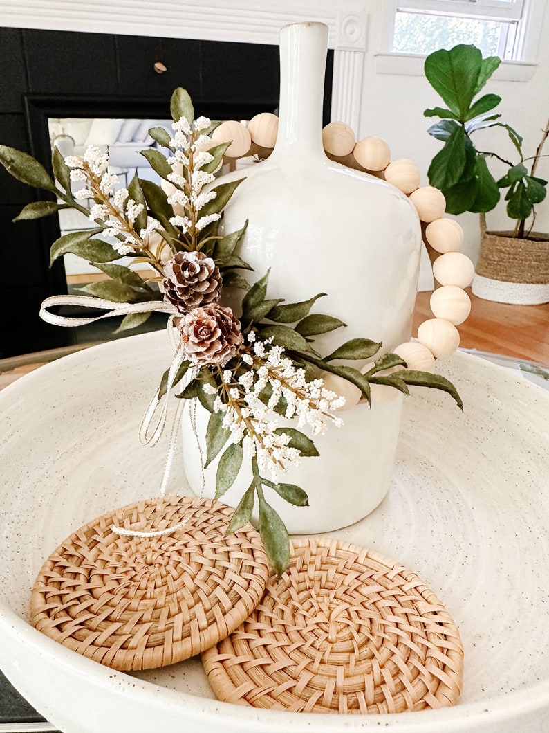 K&ouml;nnte beinhalten: Eine wei&szlig;e Keramikvase mit einem dekorativen Blumenarrangement und einer Holzperlen-Girlande. Zwei gewebte Rattan-Untersetzer liegen in einer gesprenkelten wei&szlig;en Schale. Das Arrangement umfasst gr&uuml;ne Bl&auml;tter, wei&szlig;e Beeren und braune Tannenzapfen.