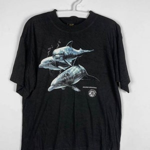 May include: Black t-shirt with a graphic of three dolphins swimming in the ocean. The text "Dolphin Migration" is printed on the bottom of the shirt. The shirt has a Planet Earth logo.