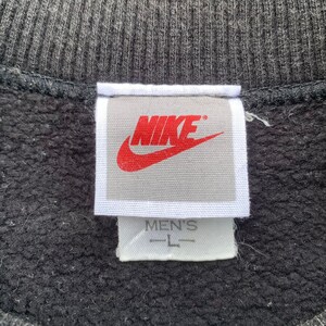 K&ouml;nnte beinhalten: Ein wei&szlig;es Etikett mit dem Wort "NIKE" in roten Buchstaben und einem roten Swoosh-Logo. Das Etikett tr&auml;gt au&szlig;erdem die Aufschrift "MEN'S" und "L" in schwarzen Buchstaben.