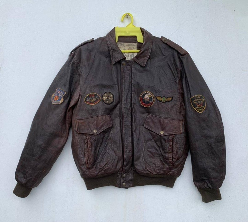 May include: A brown leather bomber jacket with multiple patches, including a US Air Force patch, a 357th Fighter Squadron patch, and a 1st Fighter Group patch.