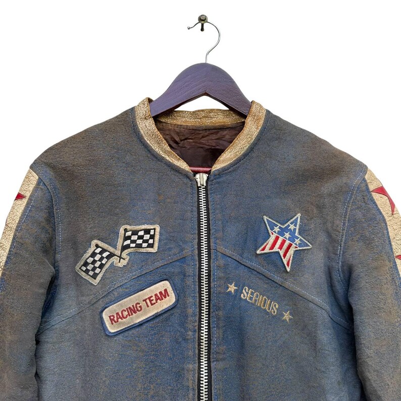 Op de afbeelding: Een blauw leren jack met een ritssluiting en verschillende patches. De patches omvatten een geruite vlag, een ster met rode, witte en blauwe strepen, en een patch met de tekst "Racing Team". Het jack heeft ook een patch met de tekst "Serious" met sterren aan beide kanten.
