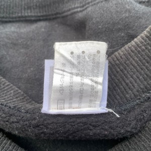 K&ouml;nnte beinhalten: Ein wei&szlig;es Etikett mit schwarzem Text in Japanisch. Das Etikett ist an einem schwarzen Kleidungsst&uuml;ck befestigt. Der Text lautet "(C) バンダイ"