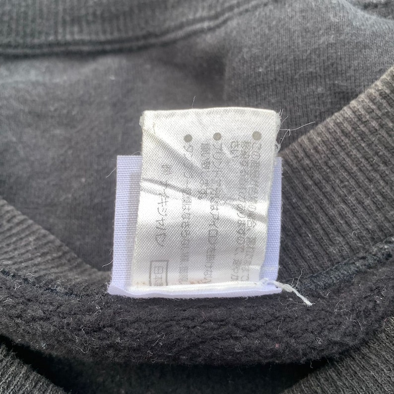 K&ouml;nnte beinhalten: Ein wei&szlig;es Etikett mit schwarzem Text in Japanisch. Das Etikett ist an einem schwarzen Kleidungsst&uuml;ck befestigt. Der Text lautet "(C) バンダイ"