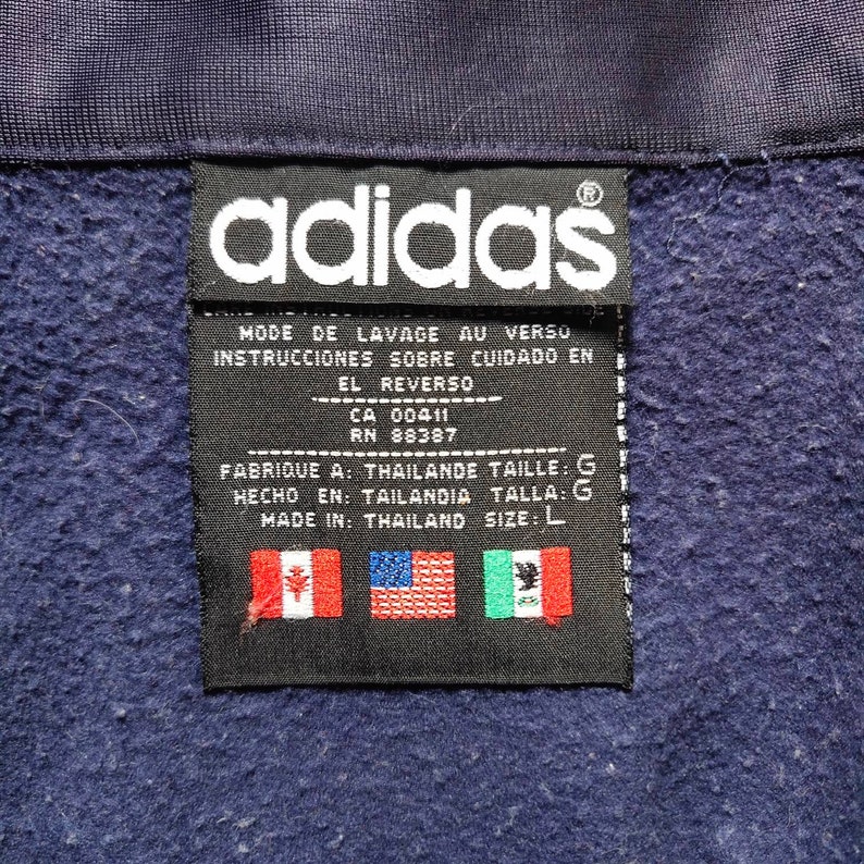 May include: Black and white adidas label with text in French, Spanish, and English. The label includes the size, L, and the country of origin, Thailand. The label also includes the Canadian, American, and Mexican flags.