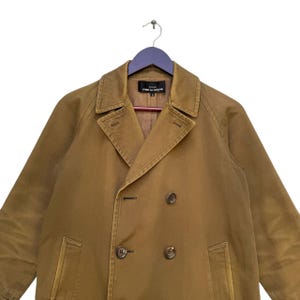 May include: A tan double-breasted coat with a notched collar and button fastenings. The coat is hanging on a purple hanger. The label reads "tricot COMME des GAR&Ccedil;ONS".