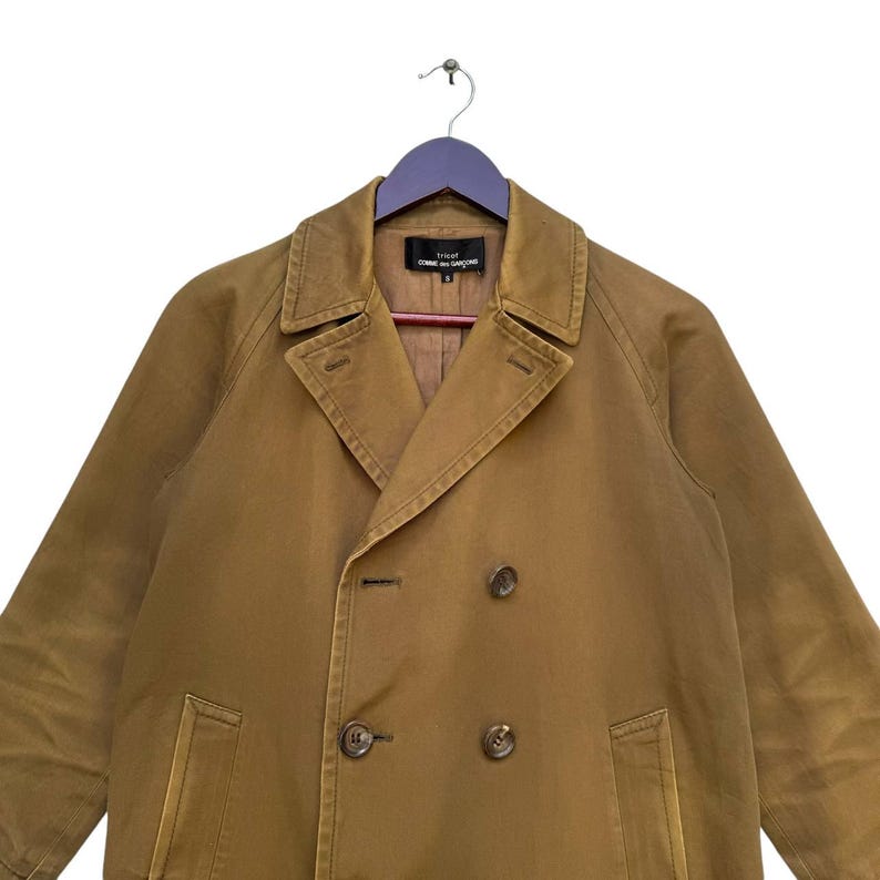 May include: A tan double-breasted coat with a notched collar and button fastenings. The coat is hanging on a purple hanger. The label reads "tricot COMME des GAR&Ccedil;ONS".