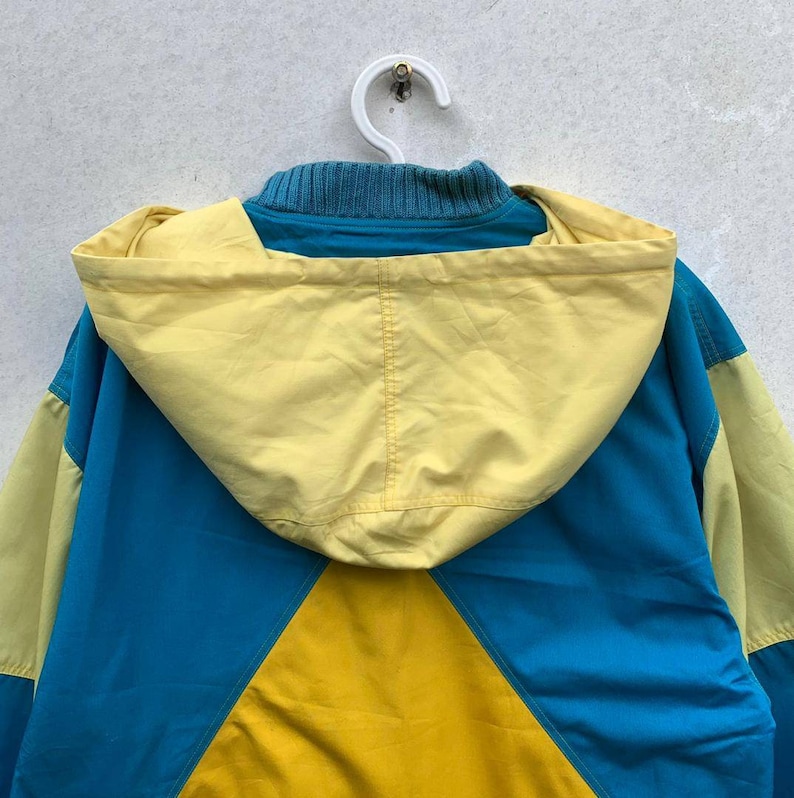 May include: A blue, yellow, and light yellow hooded jacket. The jacket has a color block design with a large yellow triangle on the front.
