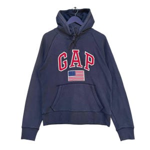 Könnte beinhalten: Ein marineblauer Kapuzenpullover mit einem gestickten "GAP"-Logo in Rot und Weiß und einer kleinen amerikanischen Flagge.