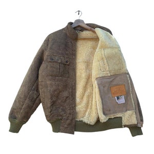 Puede incluir: Una chaqueta bomber de cuero marrón con forro de piel de oveja. La chaqueta tiene cierre de cremallera, dos bolsillos en el exterior y un parche en el interior con el texto "Real Leather".