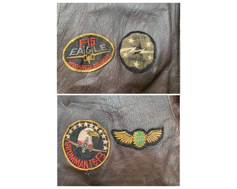 May include: A brown leather jacket with four embroidered patches. The patches are for the F-15 Eagle, McDonnell Douglas, 1st Fighter Squadron, and the US Air Force.