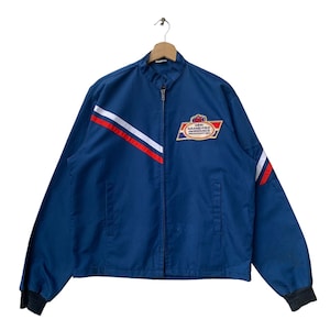 Op de afbeelding: Een blauwe vintage jas met een rood-wit-blauw streepontwerp. De jas heeft een ritssluiting en een patch op de borst met de tekst "25th GRAND PRIX MONACO".