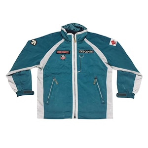 May include: Teal and white zip-up jacket with a high collar. The jacket features white accents on the sleeves and shoulders, with the text "GMC" and "DESCENTE" on the chest. A Canadian maple leaf patch is visible on the right shoulder.