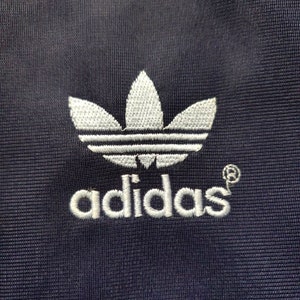 May include: A white embroidered Adidas logo on a dark blue fabric background. The logo features the iconic three-leaf clover design and the word "adidas" in white lettering.