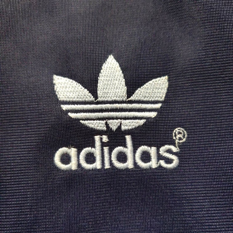May include: A white embroidered Adidas logo on a dark blue fabric background. The logo features the iconic three-leaf clover design and the word "adidas" in white lettering.