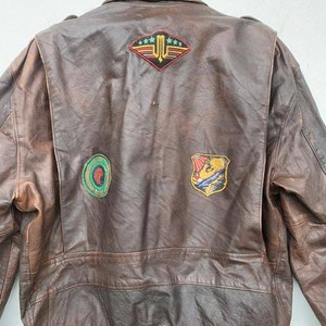 May include: Brown leather bomber jacket with multiple patches on the back. The patches feature a variety of designs, including a winged emblem with stars, a circular design with a green and red color scheme, and a shield with a colorful design.