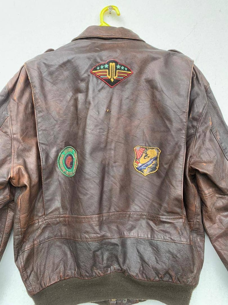 May include: Brown leather bomber jacket with multiple patches on the back. The patches feature a variety of designs, including a winged emblem with stars, a circular design with a green and red color scheme, and a shield with a colorful design.