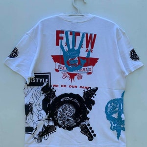 May include: White t-shirt with a graphic print of a hand with the text "FTW" and "4 year anniversary" in red and blue. The shirt also features a graphic print of a skull, guitars, and the text "We do our part".