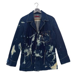 May include: A dark blue denim blazer with white paint splatter details. The jacket features a classic collar, button closure, two front pockets, and a small pocket on the chest. The blazer is hanging on a wooden hanger.