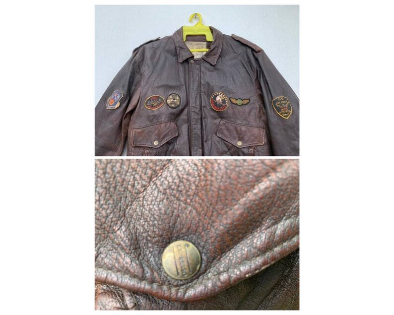 May include: A brown leather bomber jacket with multiple patches, including a U.S. Air Force patch, a  "U.S. Air Force" patch, and a "U.S. Army" patch. The jacket has a zipper closure and two pockets.