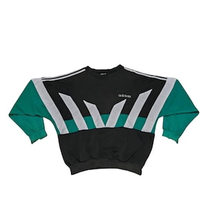 Op de afbeelding: Zwart, groen en wit Adidas sweatshirt. Het sweatshirt heeft een zwarte body met groene mouwen en witte en groene geometrische ontwerpen op de borst. Het Adidas-logo staat op de borst.