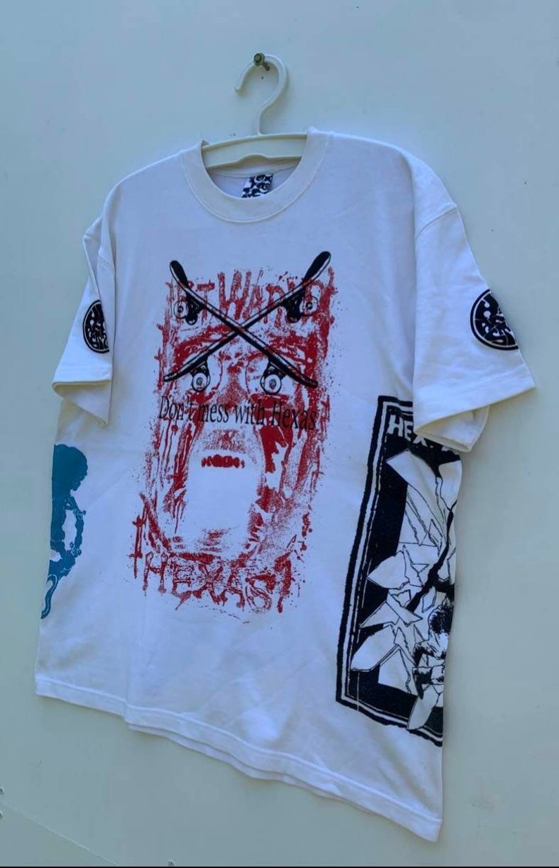 May include: White t-shirt with a graphic print of a skull with crossed skateboards and the text "Don't mess with Texas". The shirt also has a graphic print of a broken window with the text "HE" on the right side.