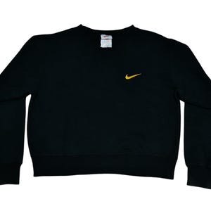 May include: Black crew neck sweatshirt with long sleeves. A small yellow Nike swoosh logo is on the left chest. The sweatshirt has a ribbed collar, cuffs, and hem. A white tag is visible at the back of the neck.