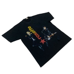 May include: Black t-shirt with a red star and the text "Roxette" in yellow and orange letters. Two people are pictured on the shirt, one holding a guitar.