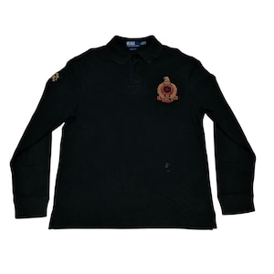 May include: Black long-sleeve polo shirt with a collared neckline and a three-button placket. Features a crest emblem on the chest and a smaller emblem on the left sleeve. The emblems have gold and red accents.