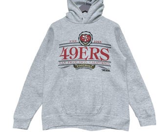 Puede incluir: Sudadera con capucha gris claro con el texto "49ERS" en rojo y blanco, con "SAN FRANCISCO, CALIFORNIA FOOTBALL" debajo. La sudadera tiene un bolsillo delantero y una capucha con cordón.