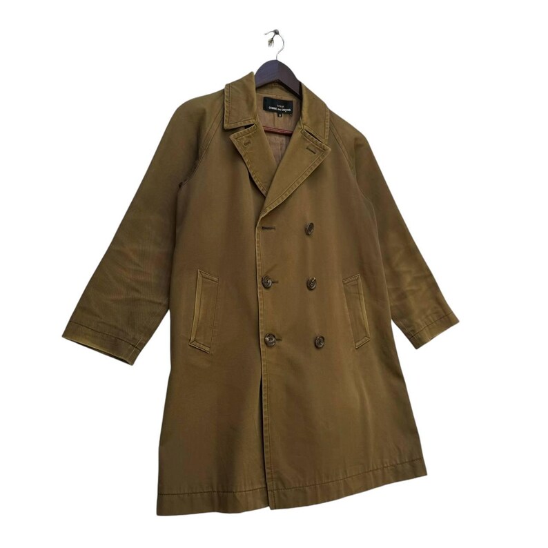 May include: An olive-green double-breasted coat with a notched collar and button fastenings. The coat features long sleeves and two front pockets. It is suspended from a wooden hanger.
