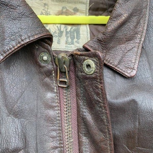 May include: Close-up of a brown leather jacket with a zipper closure. The jacket has a label that reads "Leathernecks" and "Vetement cuir et peau" with a small image of a man wearing a similar jacket. The label also says "Made in Korea".