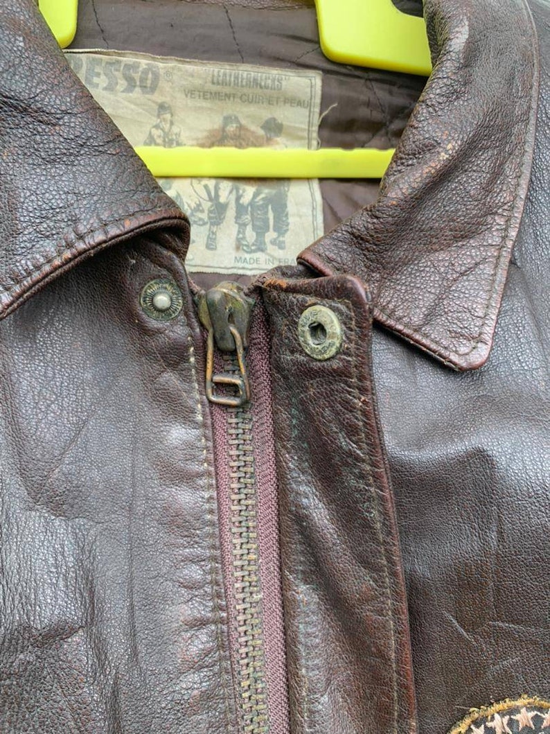 May include: Close-up of a brown leather jacket with a zipper closure. The jacket has a label that reads "Leathernecks" and "Vetement cuir et peau" with a small image of a man wearing a similar jacket. The label also says "Made in Korea".
