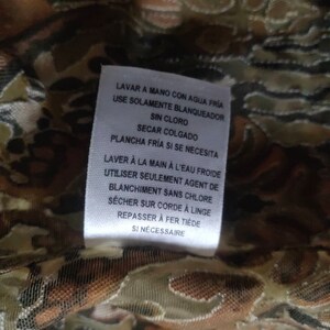 May include: White care label with instructions for washing and ironing a garment in both English and French. The label reads "Lavar a mano con agua fr&iacute;a" and "Use solamente blanqueador sin cloro" in Spanish, and "Laver &agrave; la main &agrave; l'eau froide" and "Utiliser seulement agent de blanchiment sans chlore" in French.