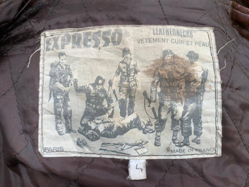 May include: A brown leather jacket with a patch that says "Expresso Leathernecks Vetement Cuir et Peau Paris Made in France". The patch features a black and white illustration of five soldiers.