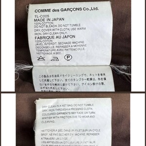 May include: Three white fabric tags with black text, attached to a brown garment. The tags provide care instructions, including "Made in Japan," "100% Cotton," and dry cleaning details. The brand name "COMME des GAR&Ccedil;ONS" is visible.