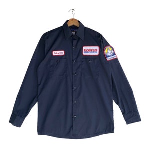 May include: A dark blue long-sleeved work shirt with two patch pockets and a button-down front. The shirt has a white name tag that says "Takahiro" and a red, white, and blue Costco Wholesale patch on the left chest. There is also a smaller patch with a cartoon character on the right chest.