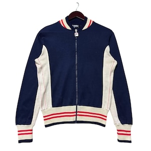Puede incluir: Chaqueta azul marino con cremallera, paneles laterales color crema y rayas rojas en el cuello, los puños y el dobladillo. La chaqueta tiene una cremallera completa y está colgada de una percha de madera. Estilo vintage.