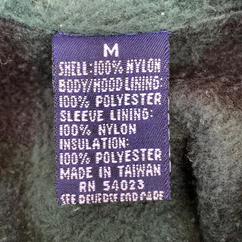 May include: A purple fabric label with white text that reads "M", "SHELL: 100% NYLON", "BODY/HOOD LINING: 100% POLYESTER", "SLEEVE LINING: 100% NYLON", "INSULATION: 100% POLYESTER", "MADE IN TAIWAN", "RN 54023", and "SEE DEUEDEF END PADE".