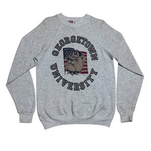 Könnte beinhalten: Ein graues Georgetown University Sweatshirt mit langen Ärmeln. Auf der Vorderseite befindet sich ein kreisförmiges Design mit den Worten "Georgetown University" und einer Bulldogge mit Hut vor einer amerikanischen Flagge.