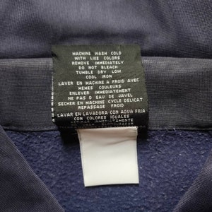 May include: A navy blue care label with washing instructions in English, French, and Spanish. The label includes instructions for machine washing, drying, and ironing. It also states to remove the garment immediately after washing and not to use bleach.