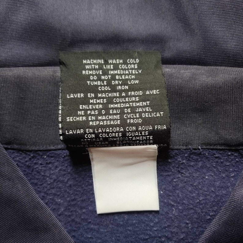 May include: A navy blue care label with washing instructions in English, French, and Spanish. The label includes instructions for machine washing, drying, and ironing. It also states to remove the garment immediately after washing and not to use bleach.