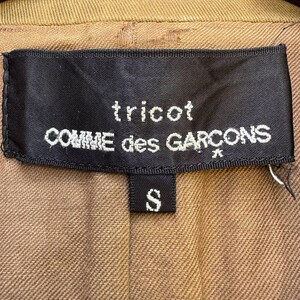 May include: A close-up shot of a black label with white text reading "tricot COMME des GAR&Ccedil;ONS" and a smaller label with the letter "S". The label is stitched onto a tan fabric.