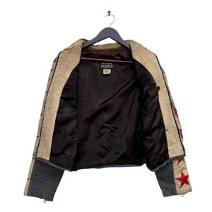 Op de afbeelding: Een beige leren jack met rode en witte sterren op de mouwen. Het jack is opengezipt en de voering is donkerbruin. Het label in het jack staat "Team Debris".