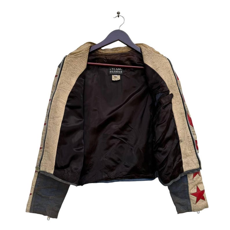 Op de afbeelding: Een beige leren jack met rode en witte sterren op de mouwen. Het jack is opengezipt en de voering is donkerbruin. Het label in het jack staat "Team Debris".