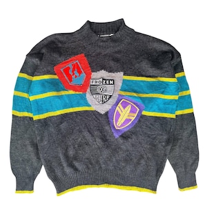 Könnte beinhalten: Dunkelgrauer Pullover mit Stehkragen. Der Pullover hat drei schildförmige Aufnäher mit Grafiken und dem Text "FROZEN SURF". Türkis- und gelb gestreift über der Brust.